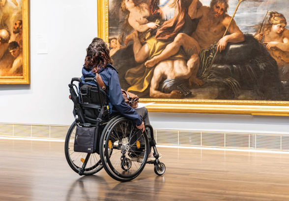 A person in a wheelchair views a large classical painting in an art gallery, surrounded by wooden floors and white walls.