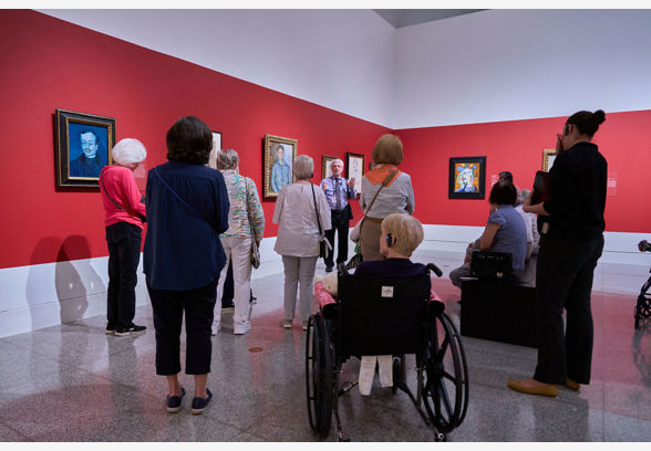 A group of older adults, including a woman in a wheelchair, gather in an art gallery to view portraits against a red wall, while a male guide discusses the artworks.